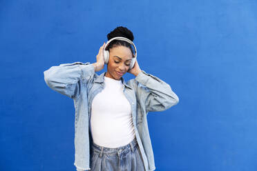 Happy woman wearing wireless headphones in front of blue wall - LMCF00855