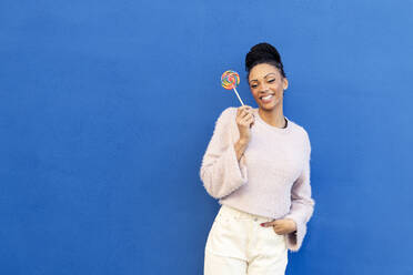 Lächelnde junge Frau, die einen Lutscher vor einer blauen Wand hält - LMCF00882