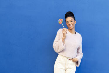 Smiling young woman holding lollipop in front of blue wall - LMCF00882