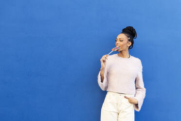 Lächelnde junge Frau isst Lutscher vor einer blauen Wand - LMCF00883