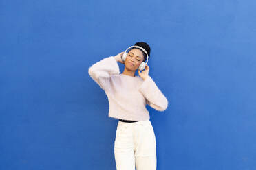 Glückliche junge Frau mit drahtlosen Kopfhörern vor einer blauen Wand - LMCF00899