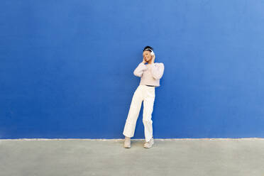 Happy woman wearing wireless headphones in front of blue wall - LMCF00901