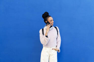 Happy young woman talking on smart phone in front of blue wall - LMCF00904