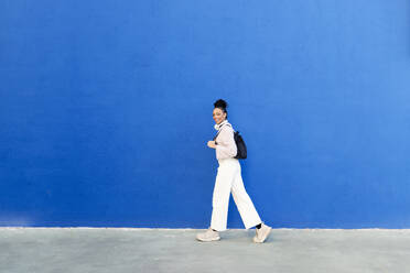 Happy woman walking in front of blue wall - LMCF00906