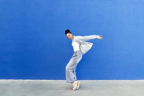 Glückliche junge Frau tanzt vor einer blauen Wand - LMCF00916