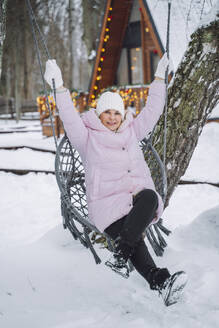 Mature woman sitting on swing near log cabin in winter - OLRF00172