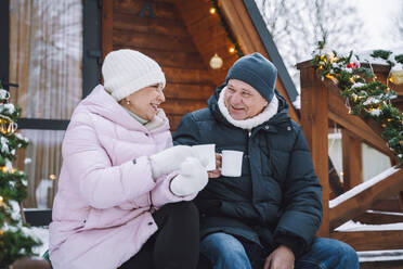 Happy mature couple enjoying hot tea sitting on porch near log cabin - OLRF00175