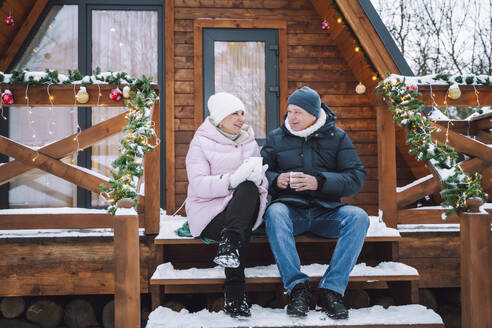 Älteres Paar sitzt und trinkt Tee auf einer Veranda in der Nähe einer Blockhütte - OLRF00176
