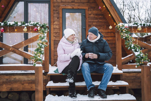 Mature couple sitting and having tea on porch near log cabin - OLRF00176