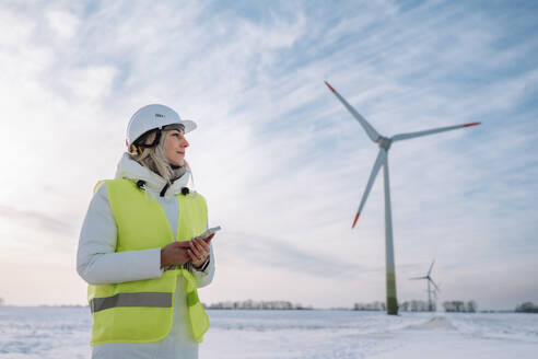 Engineer wearing hardhat holding smart phone standing at wind farm - OLRF00187