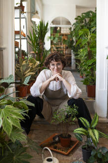 Smiling botanist leaning on elbows sitting amidst plants at nursery - VRAF00435