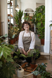 Smiling plant shop owner sitting with plants at nursery - VRAF00436