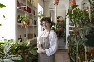 Confident botanist holding spray bottle standing amidst plants in nursery - VRAF00448