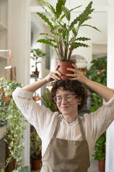 Smiling botanist with eyes closed holding plant over head in nursery - VRAF00457