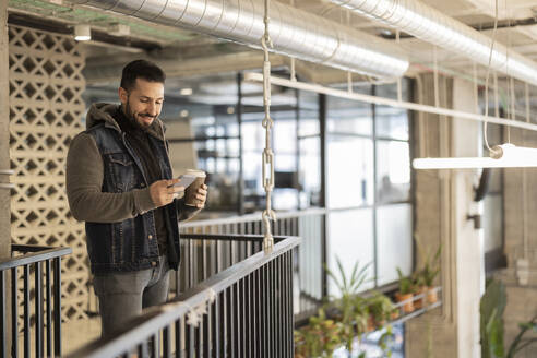 Lächelnder Geschäftsmann, der ein Smartphone benutzt und einen Einweg-Kaffeebecher im Büro hält - JCCMF11175