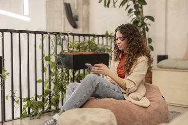 Young businesswoman sitting on bean bag using smart phone at workplace - JCCMF11176