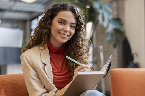 Lächelnde junge schöne Geschäftsfrau mit Tagebuch und Stift sitzt im Büro - JCCMF11184