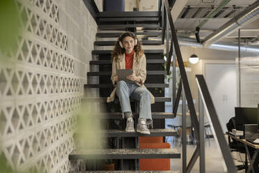 Smiling businesswoman listening to music sitting with tablet PC on staircase at office - JCCMF11206