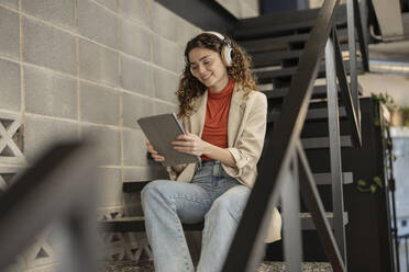 Lächelnde junge Geschäftsfrau, die mit einem Tablet-PC auf einer Treppe im Büro sitzt und Musik hört - JCCMF11207