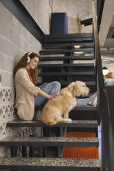 Lächelnde Geschäftsfrau, die mit ihrem Hund auf einer Treppe im Büro sitzt und Musik hört - JCCMF11209