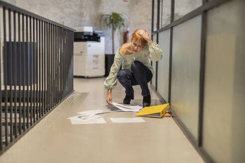 Unvorsichtige reife Geschäftsfrau, die verstreute Dokumente im Korridor eines Büros aufsammelt - JCCMF11214