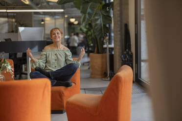 Lächelnde reife Geschäftsfrau sitzt im Schneidersitz auf einem orangefarbenen Stuhl im Büro - JCCMF11219
