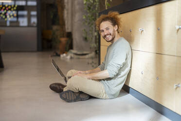 Lächelnder Geschäftsmann mit Laptop im Schneidersitz auf dem Boden sitzend am Arbeitsplatz - JCCMF11222
