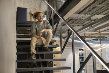 Lächelnder Geschäftsmann sitzt auf einer Treppe im Büro - JCCMF11227