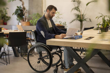 Selbstbewusster Geschäftsmann im Rollstuhl mit einer Behinderung prüft ein Dokument am Schreibtisch - JCCMF11234