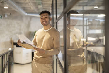 Lächelnder junger Geschäftsmann mit Dokumenten, die an einer Glaswand im Büro lehnen - JCCMF11240