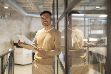 Smiling young businessman with documents leaning on glass wall at office - JCCMF11240