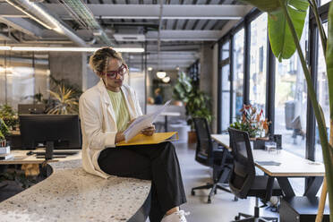 Young businesswoman reading documents sitting at workplace - JCCMF11247