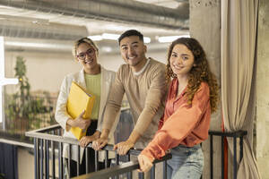Smiling young business colleagues leaning on railing at office - JCCMF11266