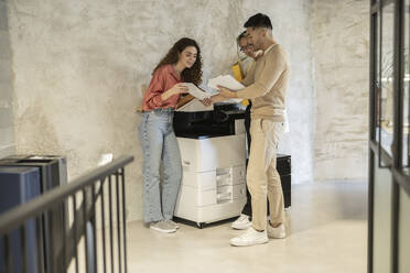 Young business colleagues discussing over documents standing by photocopier at office - JCCMF11269
