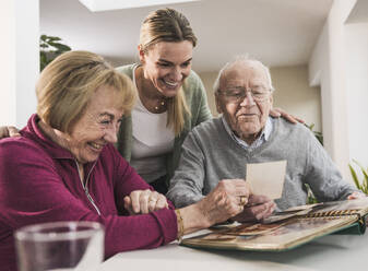 Happy home caregiver with senior couple looking at photographs - UUF31167