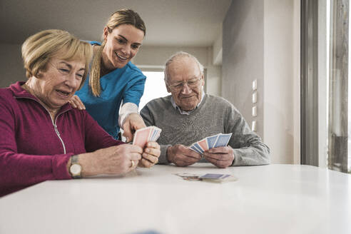 Happy senior couple playing cards with home caregiver at table - UUF31196