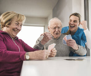 Happy home caregiver with man and woman playing cards - UUF31197