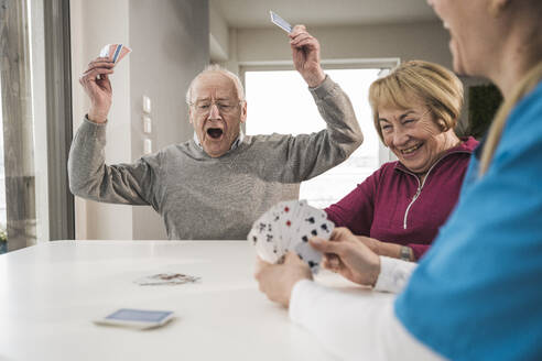 Cheerful man playing cards with woman and home caregiver at table - UUF31198