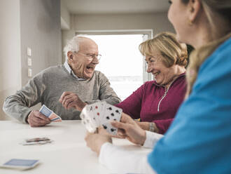 Fröhliches Paar spielt Karten mit Hauspflegerin am Tisch - UUF31199