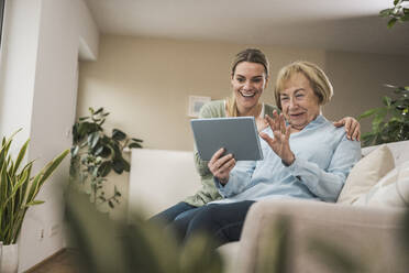 Glückliche Hauspflegerin mit älterer Frau, die im Wohnzimmer einen Tablet-PC benutzt - UUF31207