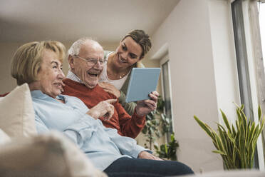 Smiling senior couple using tablet PC with home care giver in living room - UUF31211