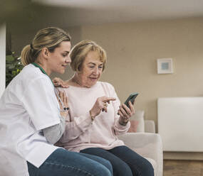 Lächelnder Arzt mit älterer Frau, die zu Hause ein Smartphone benutzt - UUF31230
