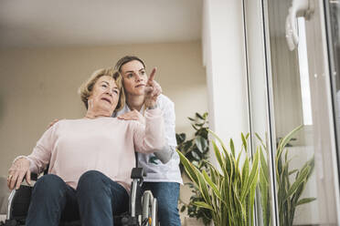Krankenschwester mit älterer Frau, die zu Hause im Rollstuhl sitzt und auf etwas zeigt - UUF31243