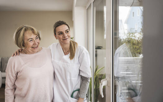 Lächelnde Krankenschwester mit älterer Frau, die durch ein Fenster im Haus schaut - UUF31247