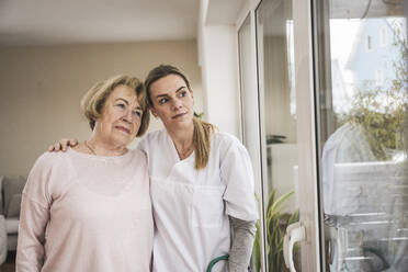 Krankenschwester mit älterer Frau, die durch ein Fenster zu Hause schaut - UUF31248