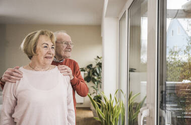 Glücklicher älterer Mann und ältere Frau schauen durch das Fenster zu Hause - UUF31250