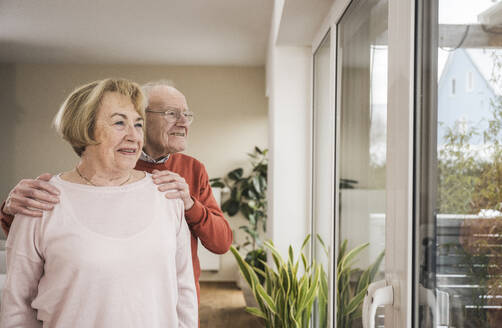 Happy senior man and senior woman looking through window at home - UUF31250