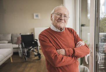 Lächelnder älterer Mann, der mit verschränkten Armen zu Hause steht - UUF31254