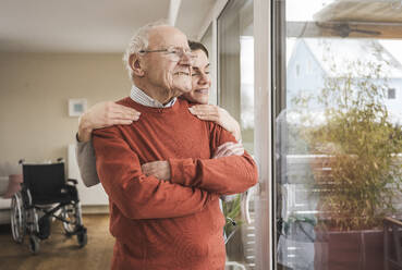 Lächelnde Hauspflegerin umarmt älteren Mann, der durch das Fenster schaut - UUF31258