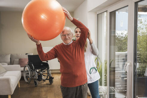 Hauspflegerin mit älterem Mann, der Übungen macht und einen Fitnessball hält - UUF31259